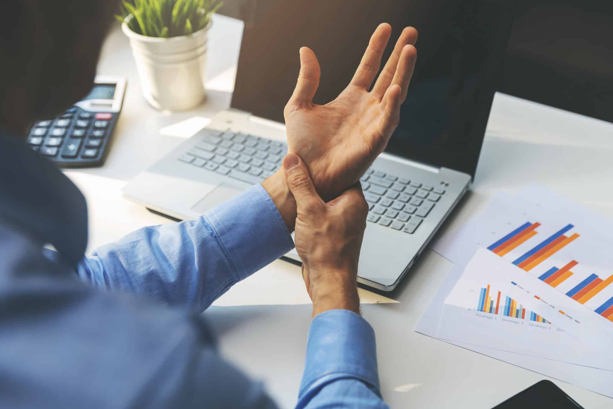 Man suffering from carpal tunnel syndrome. He's holding his wrist in pain, while sitting at desk in front of a laptop.