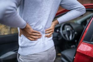 Man standing next to car holding lower back in pain from possible lumbar fracture in Dallas