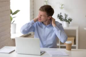 Man sitting at desk holding back of neck in pain due to spinal stenosis in Dallas, TX